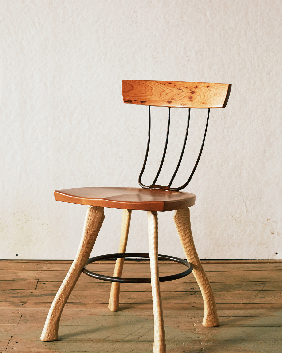 Front three-quarter view of the Bradford Woodworking Pitchfork Chair at dining height. Solid cherry seat and backrest with forged steel pitchfork tine spindles. Ax handle lathe-turned ash legs splay outward with a steel hoop stretcher connecting all four legs.