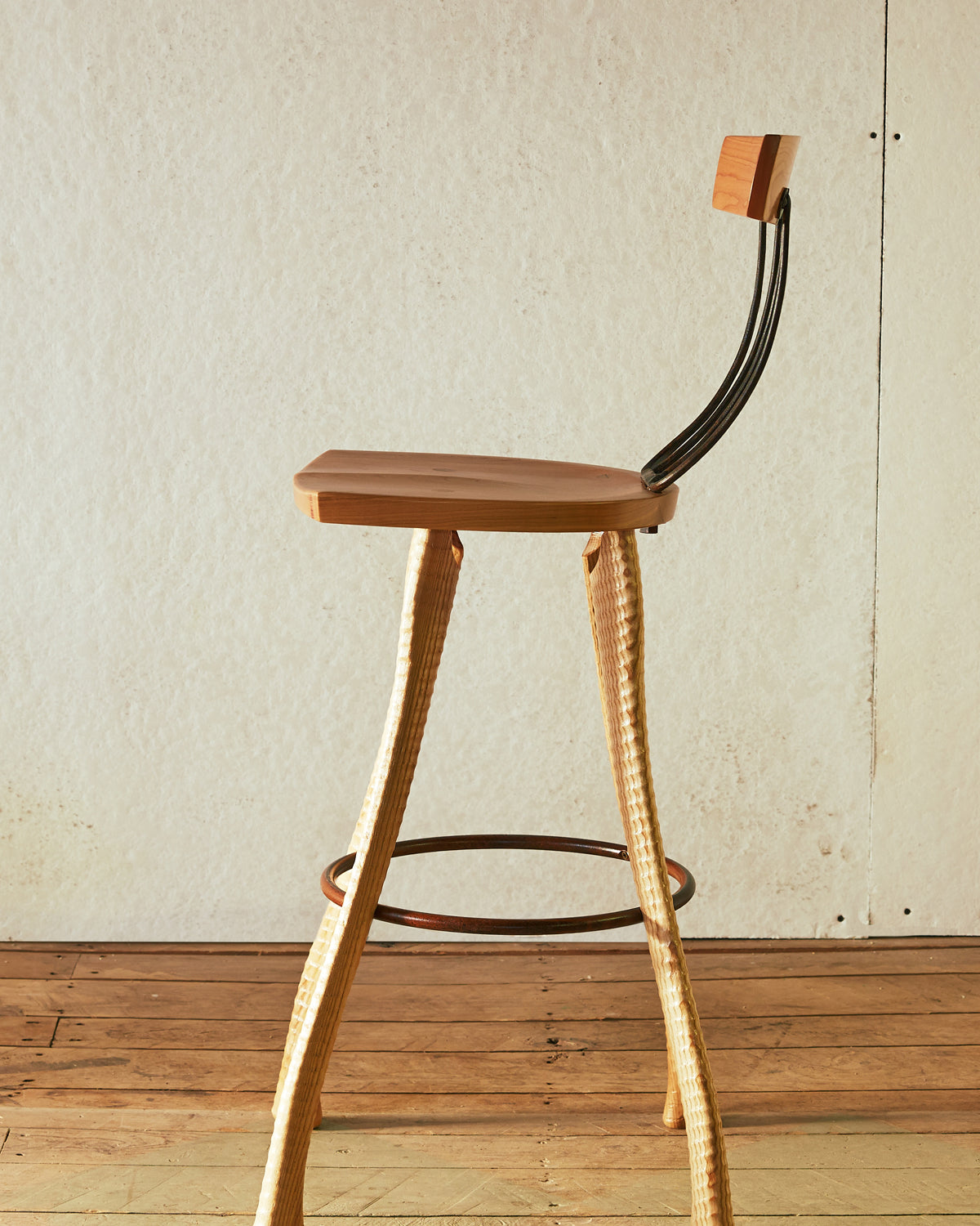 Side profile of the 25" counter height Pitchfork Chair showing the curve of the forged pitchfork tines connecting seat to backrest, ax handle lathe-turned ash legs, and steel hoop footrest. Solid cherry seat visible from the side.