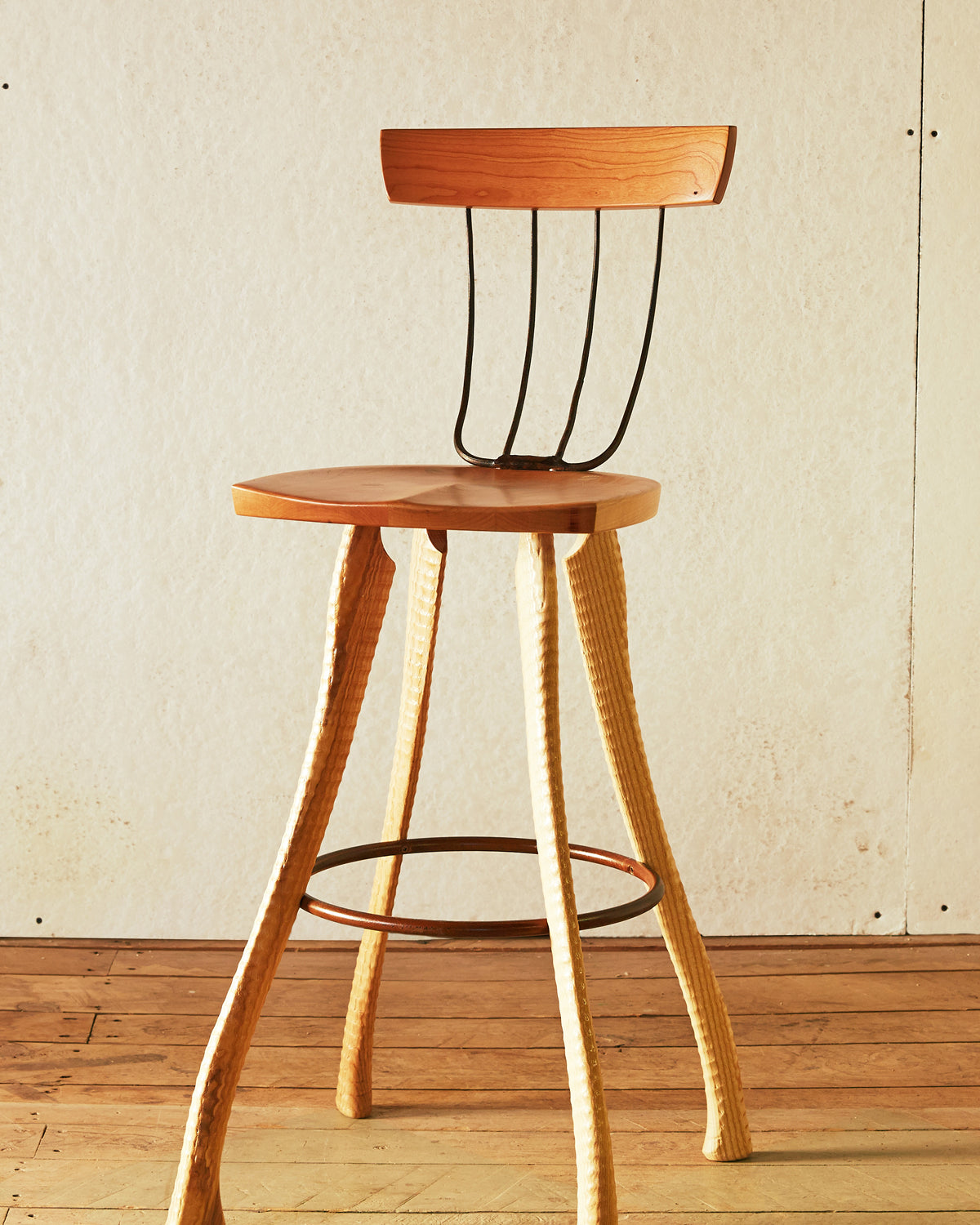 Front view of the Bradford Woodworking Pitchfork Chair at counter height. Solid cherry round seat and backrest, four forged steel pitchfork tine spindles, ax handle lathe-turned ash legs splaying outward, and a steel hoop stretcher at footrest height.