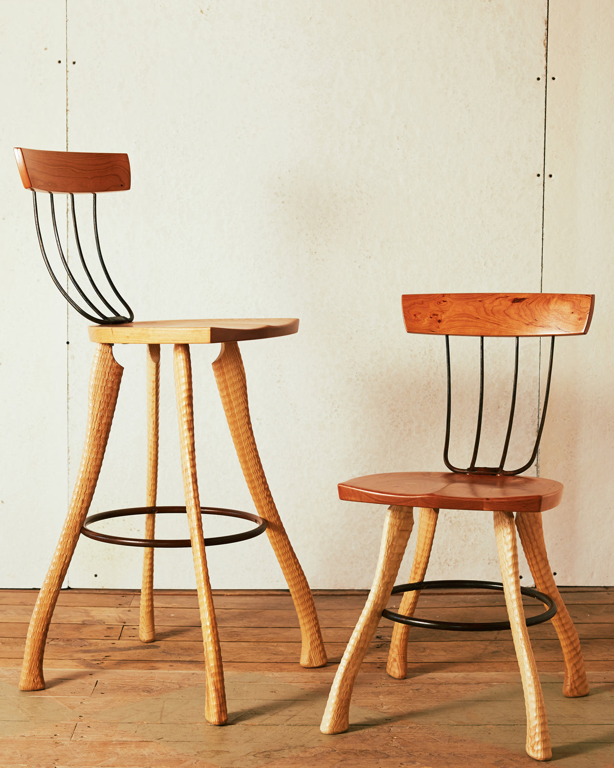 A 30" bar height and 18" dining height Bradford Woodworking Pitchfork Chair photographed together on a wood floor. Both feature solid cherry seats and backrests, forged pitchfork tine back spindles, and ax handle lathe-turned ash legs with a steel hoop stretcher and footrest.
