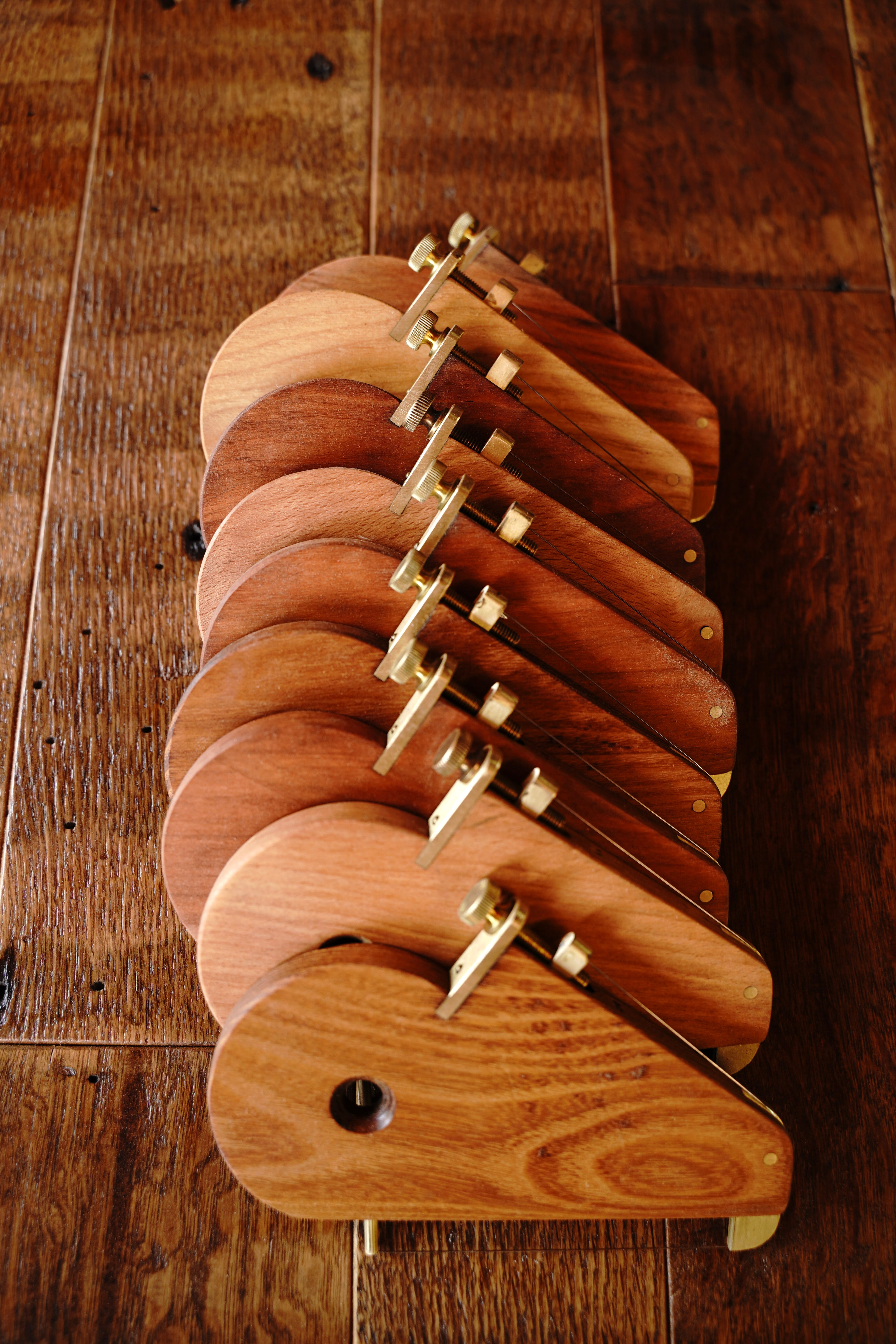 A stack of handcrafted hardwood cheese slicers with brass wire-tension hardware, arranged in a fanned display on a rustic wood surface. Each slicer features a rounded wooden paddle in varied warm wood tones, made by Bradford Woodworking.