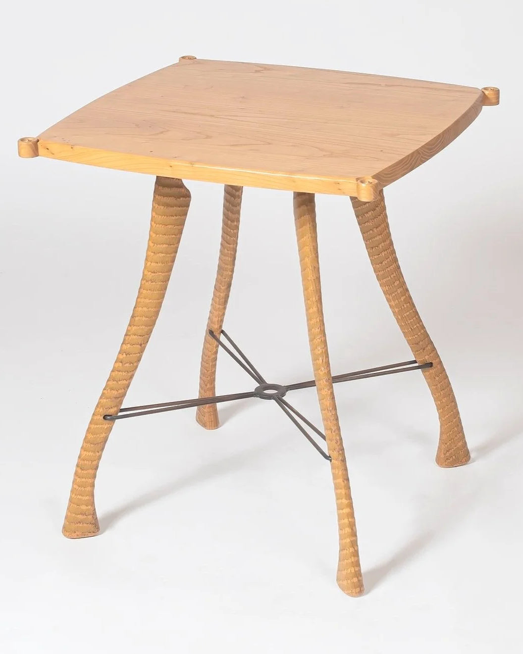 Handcrafted side table with a solid American elm top featuring clipped corners and small turned corner pegs. Four hand-textured elm legs splay outward with a ringed surface pattern running the full length of each leg, tapering to wide feet. A dark steel cross stretcher connects all four legs at a central welded ring. Photographed against a white background.