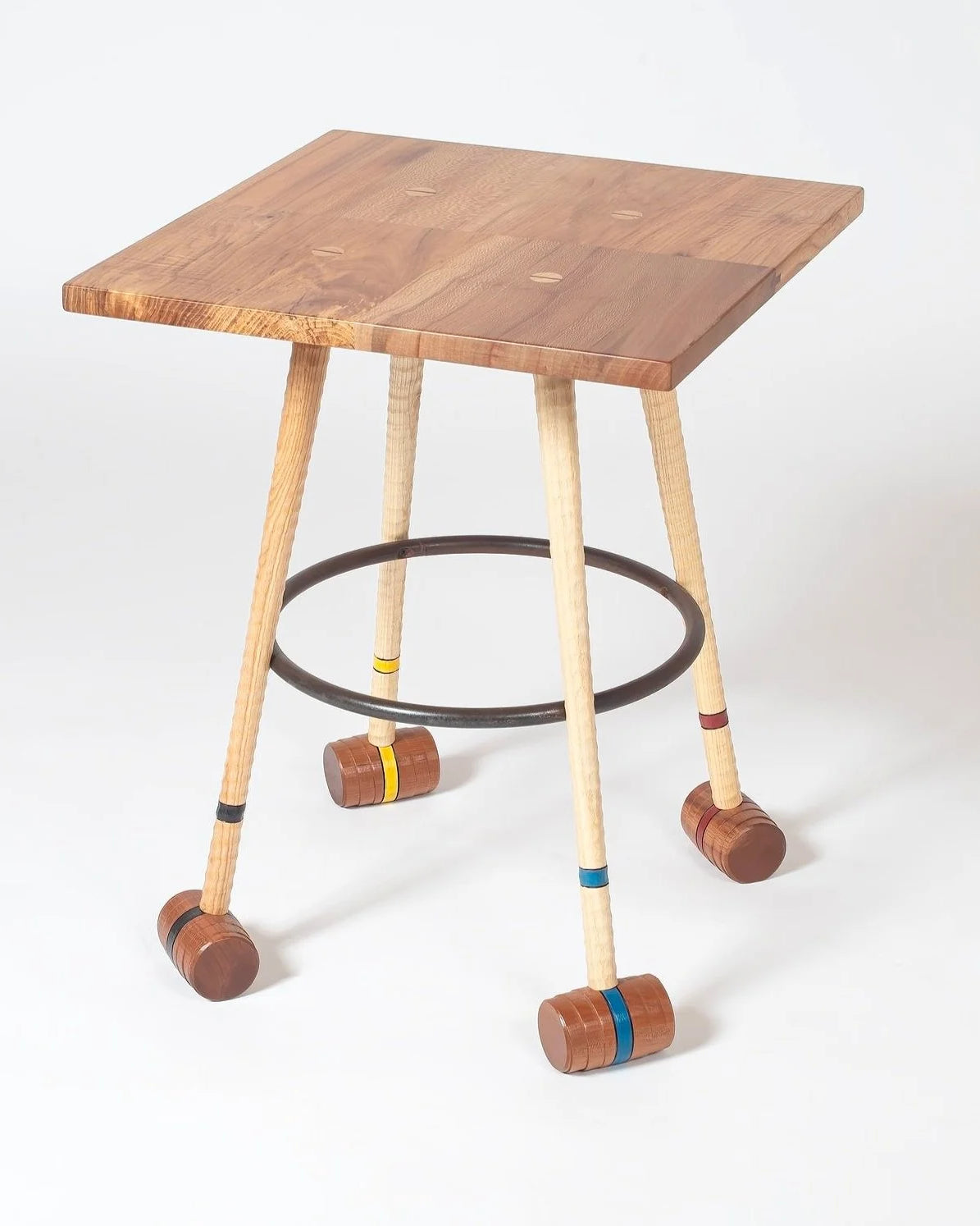 Handcrafted wooden side table with four croquet mallet legs in ash and walnut, each banded in yellow, blue, red, and black. A circular steel hoop connects the legs as a stretcher. The square tabletop is solid mixed hardwoods with natural grain variation. Shot against a white background.