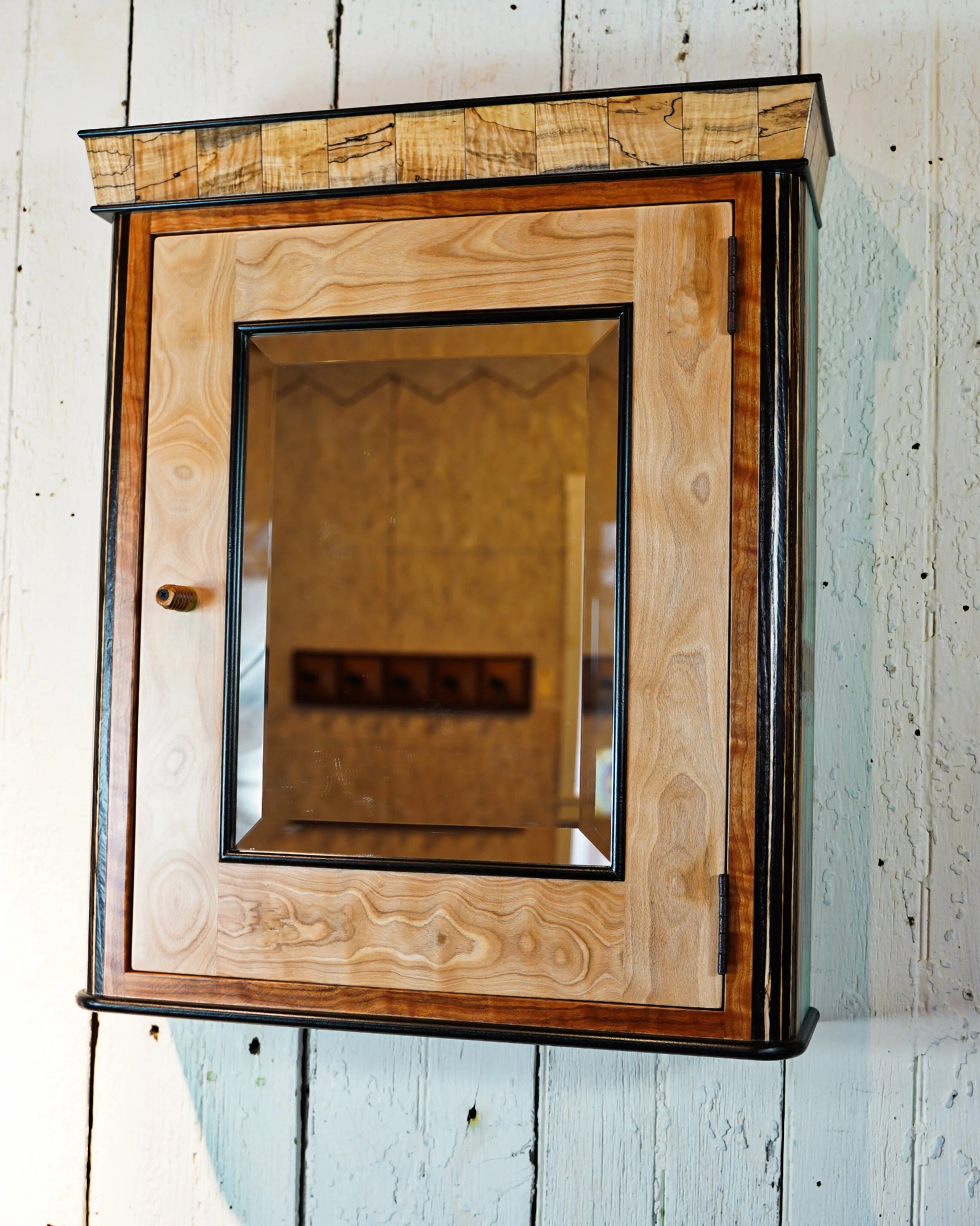 Handcrafted wall-hung medicine cabinet with a spalted maple crown showing dark fungal figure, a beech door with cherry trim, and a beveled mirror set in a black frame. A small turned wooden knob sits on the left side of the door. Cabinet is mounted on a white painted wood plank wall.