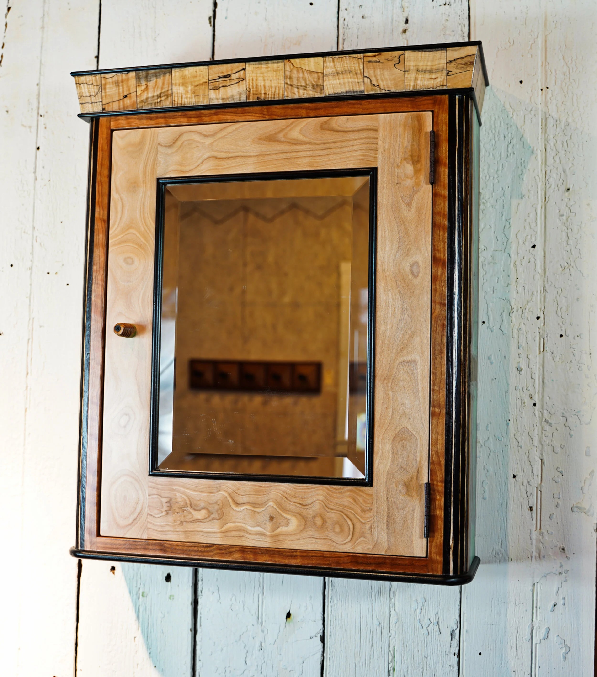 Handcrafted wall-hung medicine cabinet with a spalted maple crown showing dark fungal figure, a beech door with cherry trim, and a beveled mirror set in a black frame. A small turned wooden knob sits on the left side of the door. Cabinet is mounted on a white painted wood plank wall.
