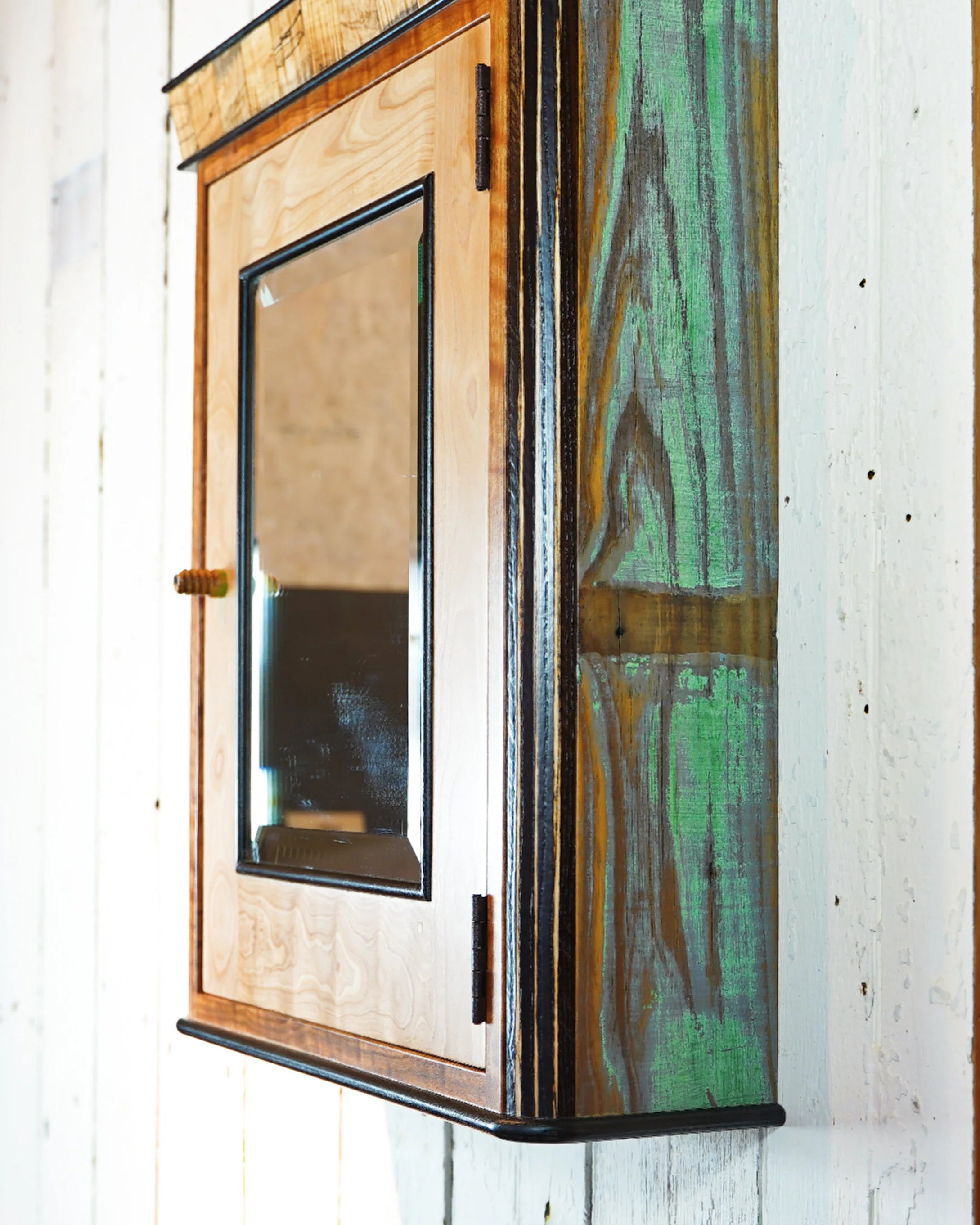Side profile of handcrafted medicine cabinet showing reclaimed barn wood panels with natural green and amber weathered color variation. Spalted maple crown is visible at the top, with black-painted trim edges running the full perimeter of the cabinet.