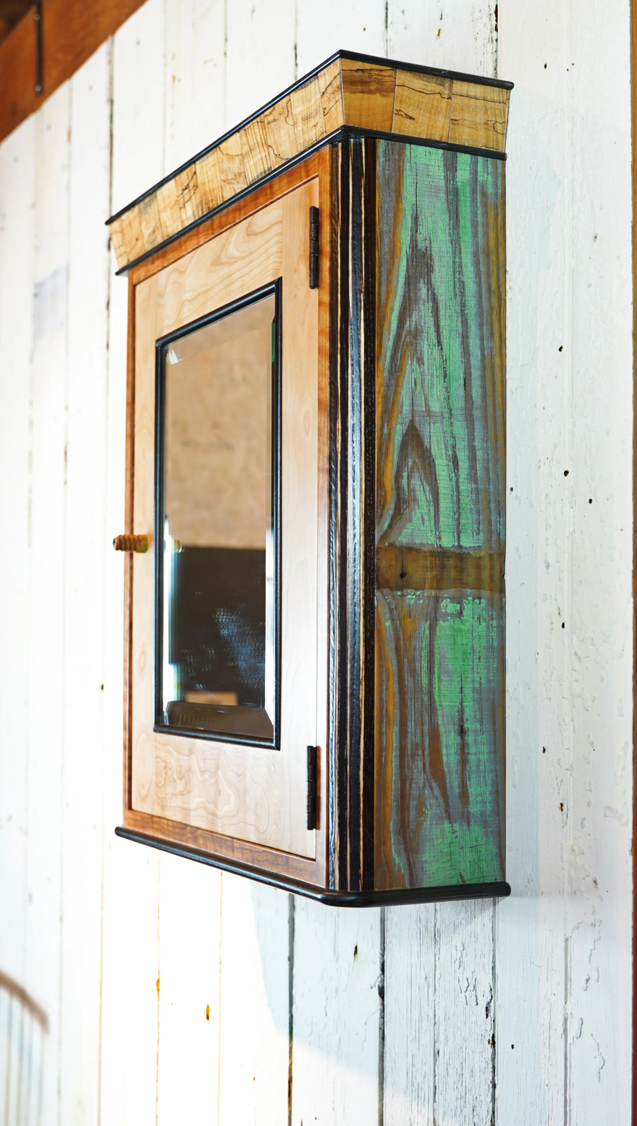 Side profile of handcrafted medicine cabinet showing reclaimed barn wood panels with natural green and amber weathered color variation. Spalted maple crown is visible at the top, with black-painted trim edges running the full perimeter of the cabinet.