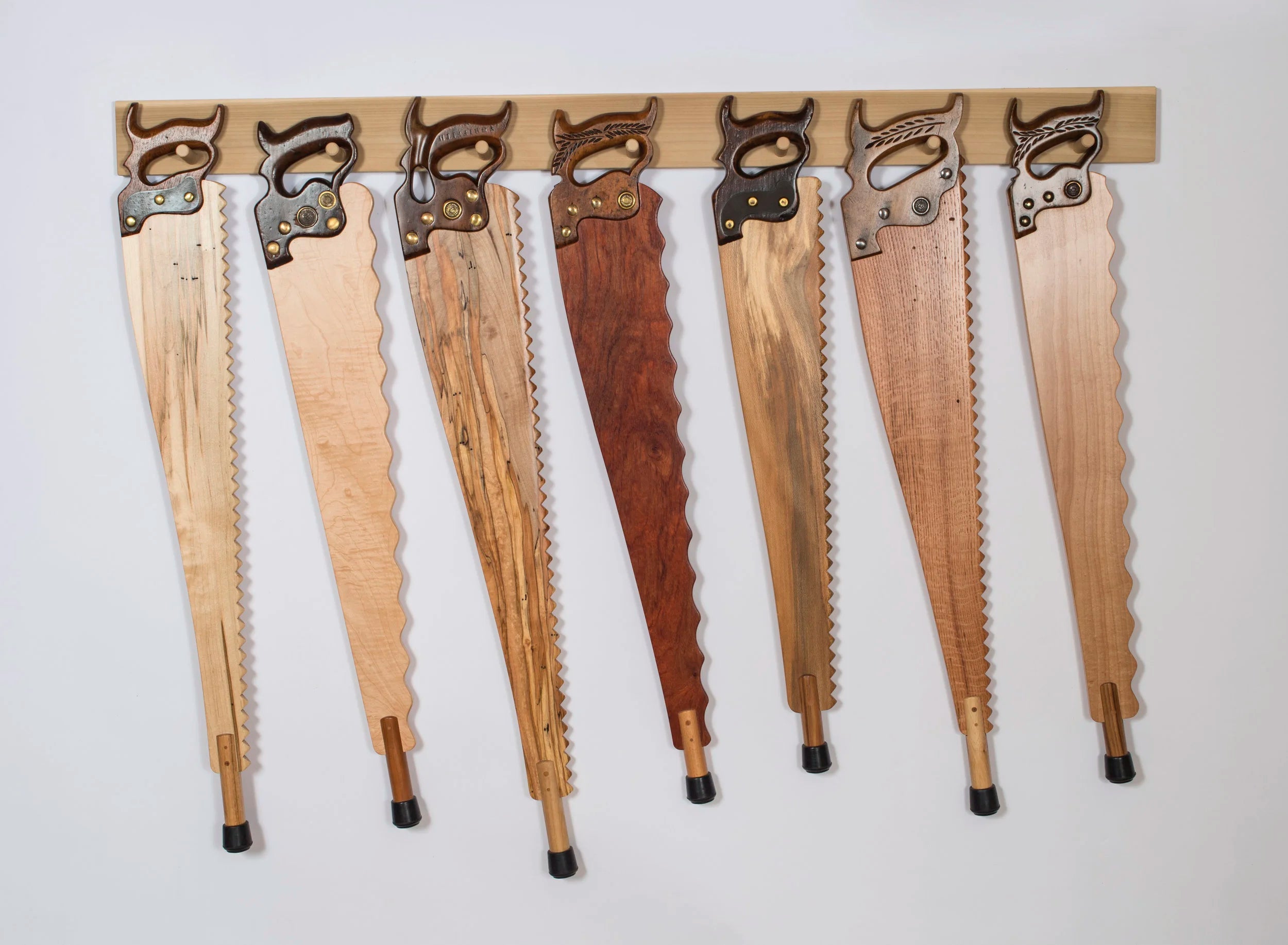 Seven handcrafted walking canes displayed on a wooden wall mount against a white background. Each cane is made from a salvaged antique hand saw — the saw blade forms the upper body of the cane, fitted with original cast hardware and brass details. The lower shafts are turned hardwood in a range of species including maple, spalted maple, walnut, and oak, each tipped with a black rubber foot.