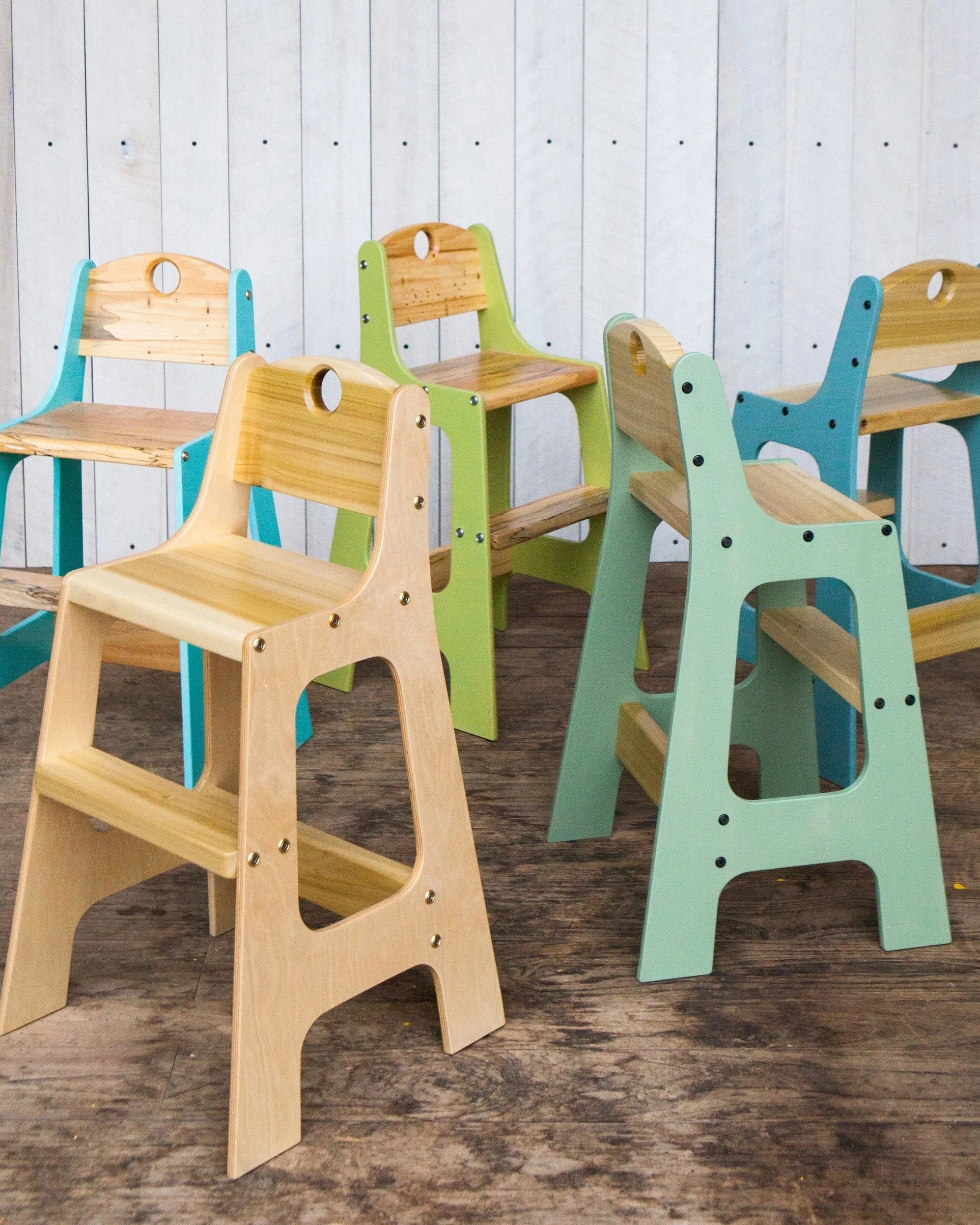 Five handcrafted toddler chairs arranged on a wood floor in front of a white plank wall. Chairs feature solid wood seats and backrests with Baltic birch plywood frames in natural, teal, green, and sage painted finishes. Each chair has visible bolt hardware and a cutout handle on the backrest.