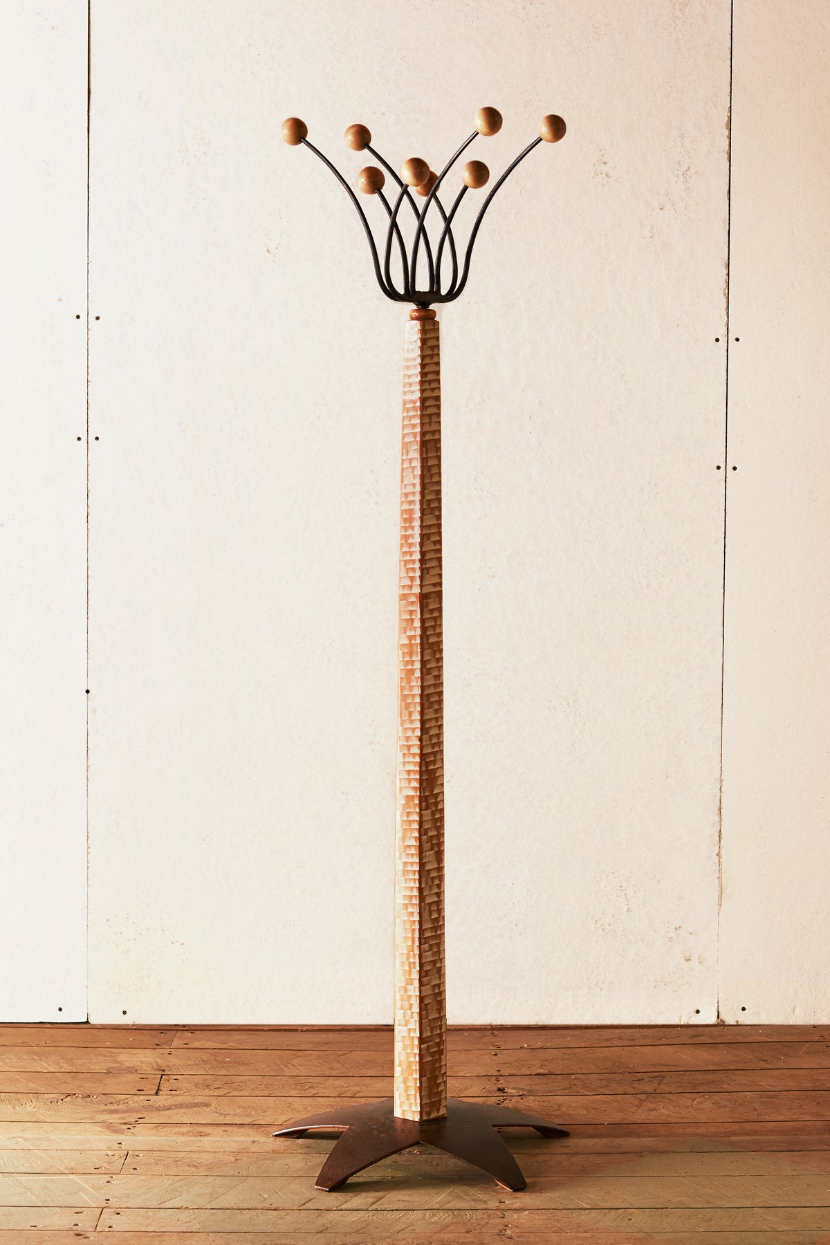 Full-length view of a Bradford Woodworking handcrafted standing coat tree. A tall whitewashed lathe-turned post supports eight forged steel arms that fan outward at the top, each tipped with a small turned wood ball hook. The post sits in a solid steel star-shaped base. Photographed against a white plank wall on a wood floor.