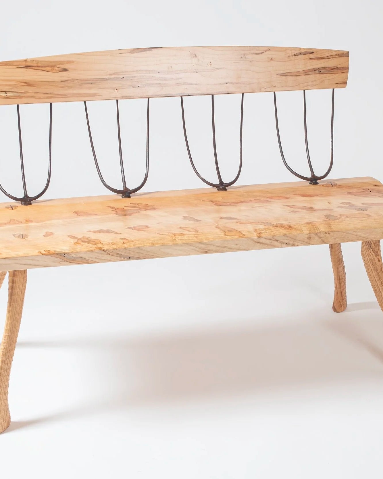 Handcrafted bench made from ambrosia maple with natural figuring and warm tonal variation. Four forged pitchfork heads serve as the back spindles, connecting the live-edge seat to a curved maple backrest. Legs are lathe-turned from ax handles with a natural outward splay. Photographed against a white background.