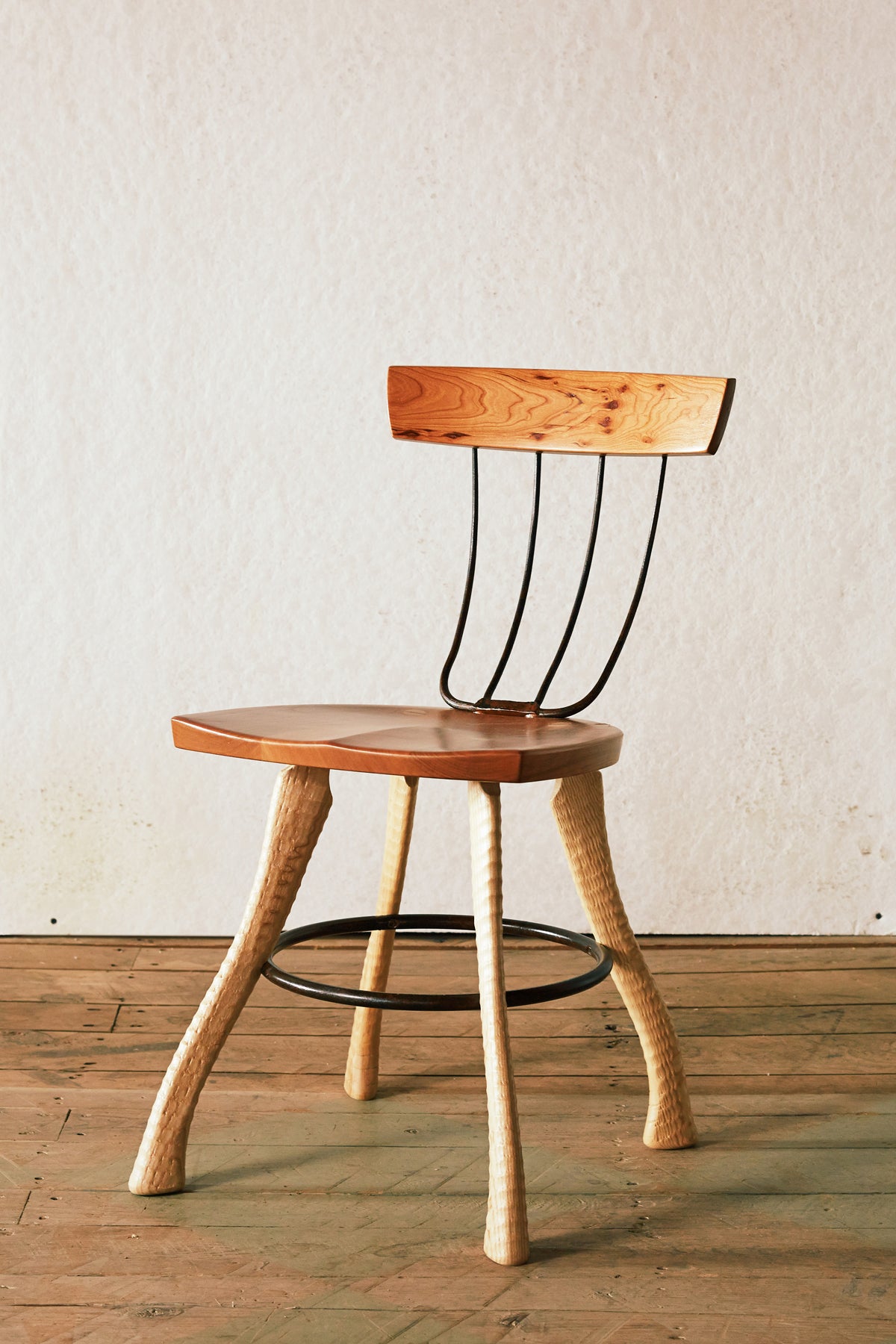Front three-quarter view of the Bradford Woodworking Pitchfork Chair at dining height. Solid cherry seat and backrest with forged steel pitchfork tine spindles. Ax handle lathe-turned ash legs splay outward with a steel hoop stretcher connecting all four legs.