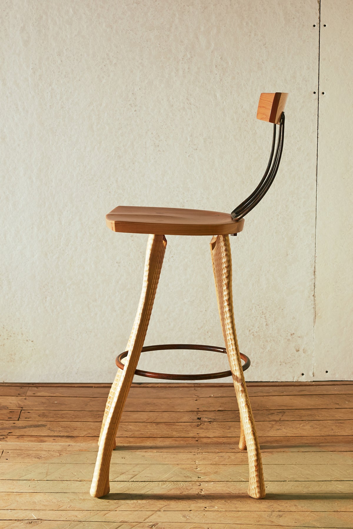 Side profile of the 25" counter height Pitchfork Chair showing the curve of the forged pitchfork tines connecting seat to backrest, ax handle lathe-turned ash legs, and steel hoop footrest. Solid cherry seat visible from the side.