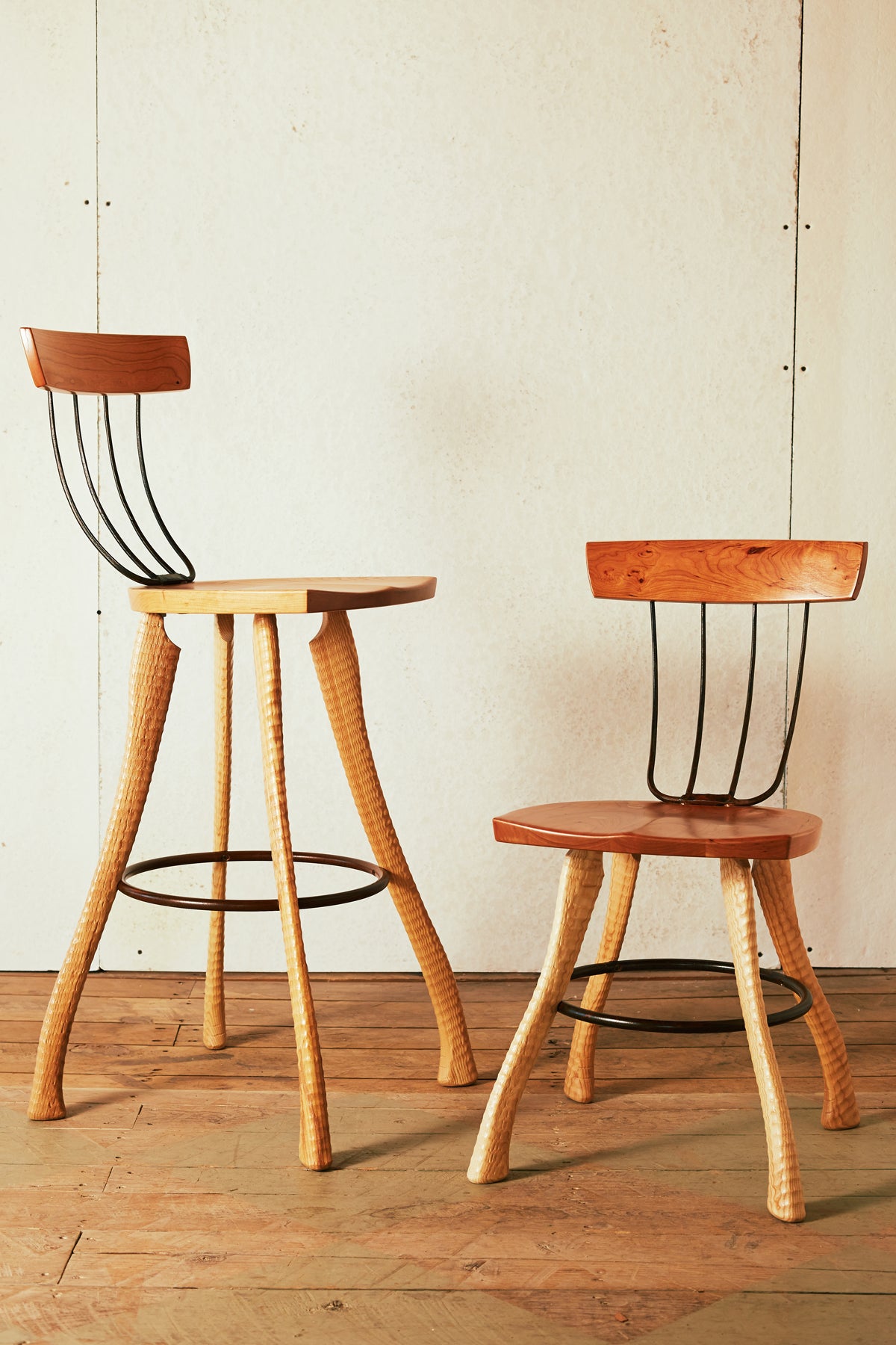 A 30" bar height and 18" dining height Bradford Woodworking Pitchfork Chair photographed together on a wood floor. Both feature solid cherry seats and backrests, forged pitchfork tine back spindles, and ax handle lathe-turned ash legs with a steel hoop stretcher and footrest.