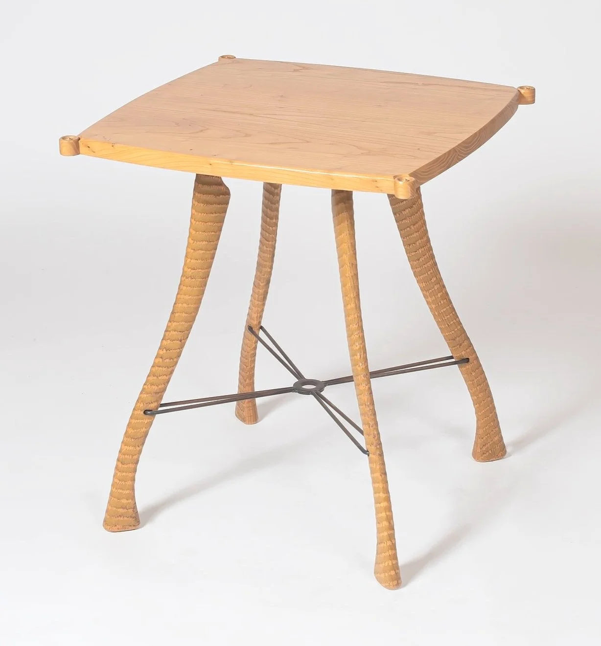 Handcrafted side table with a solid American elm top featuring clipped corners and small turned corner pegs. Four hand-textured elm legs splay outward with a ringed surface pattern running the full length of each leg, tapering to wide feet. A dark steel cross stretcher connects all four legs at a central welded ring. Photographed against a white background.
