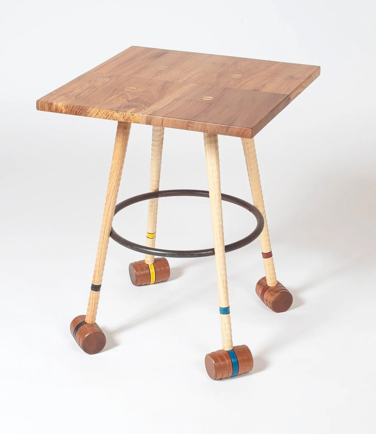 Handcrafted wooden side table with four croquet mallet legs in ash and walnut, each banded in yellow, blue, red, and black. A circular steel hoop connects the legs as a stretcher. The square tabletop is solid mixed hardwoods with natural grain variation. Shot against a white background.