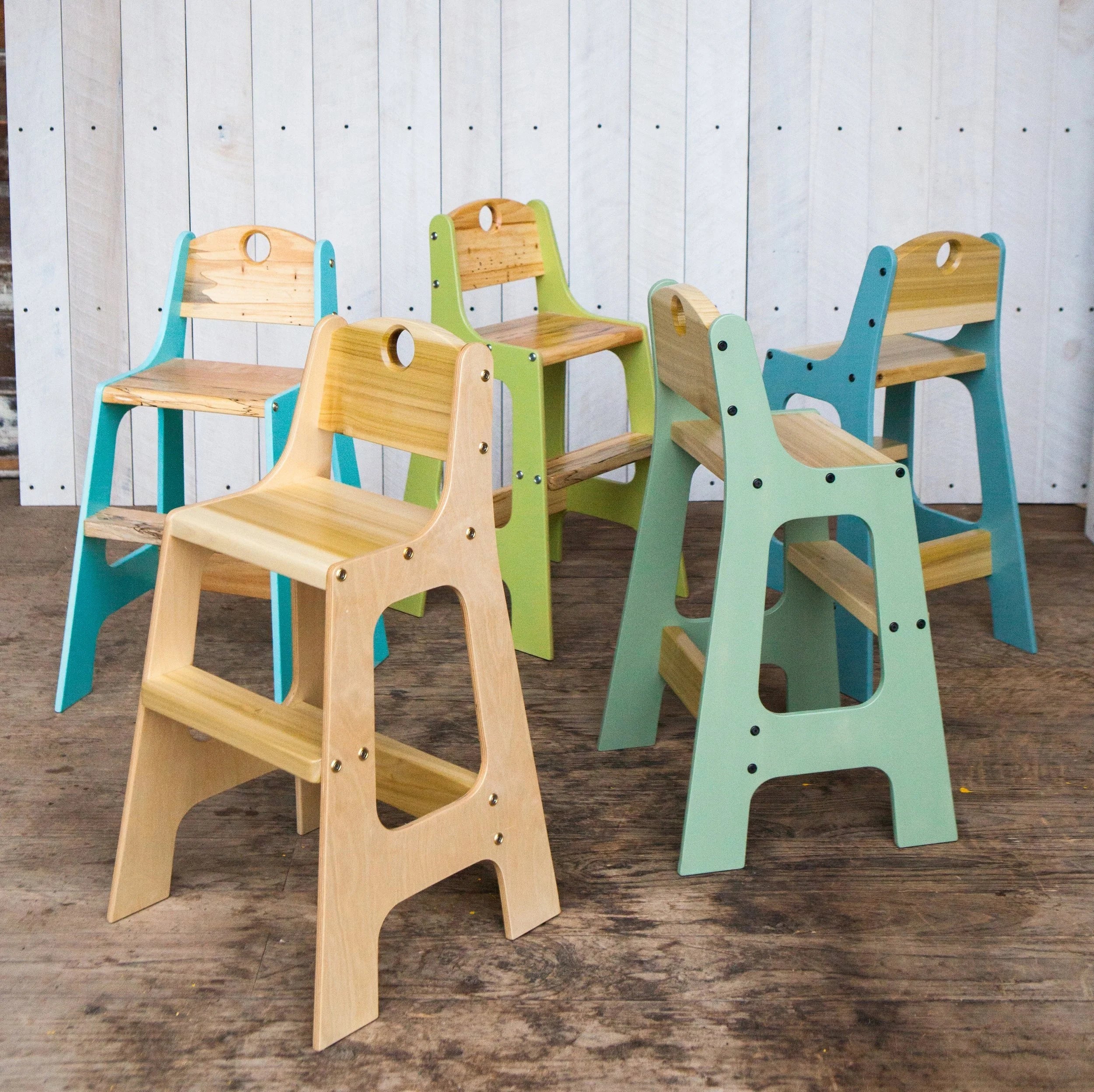 Five handcrafted toddler chairs arranged on a wood floor in front of a white plank wall. Chairs feature solid wood seats and backrests with Baltic birch plywood frames in natural, teal, green, and sage painted finishes. Each chair has visible bolt hardware and a cutout handle on the backrest.