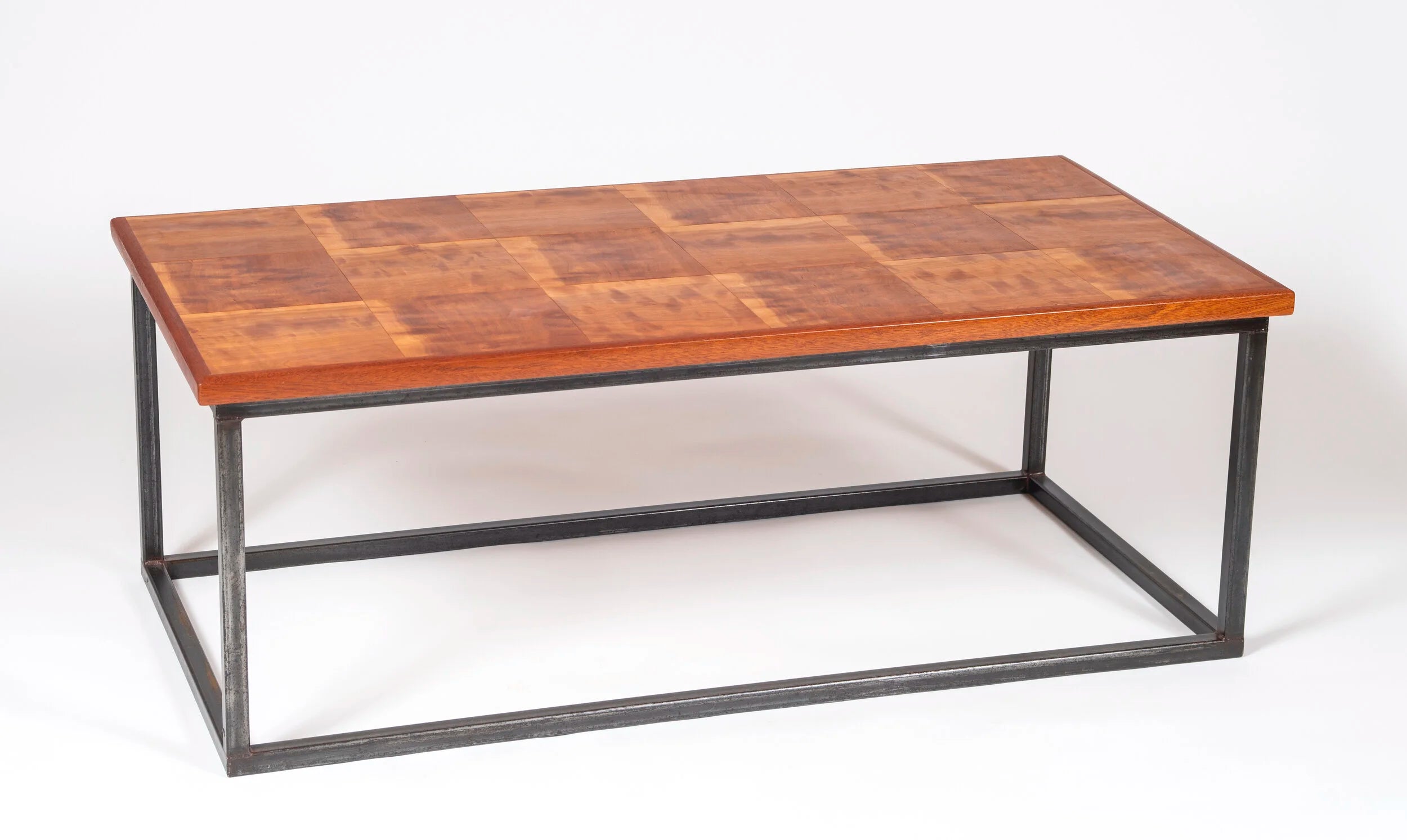 Handcrafted coffee table with a highly figured solid cherry top laid in a tiled panel pattern, showing warm amber and reddish brown tones with natural variation across each section. The base is a clean rectangular welded steel box frame in a dark finish. Photographed against a white background.