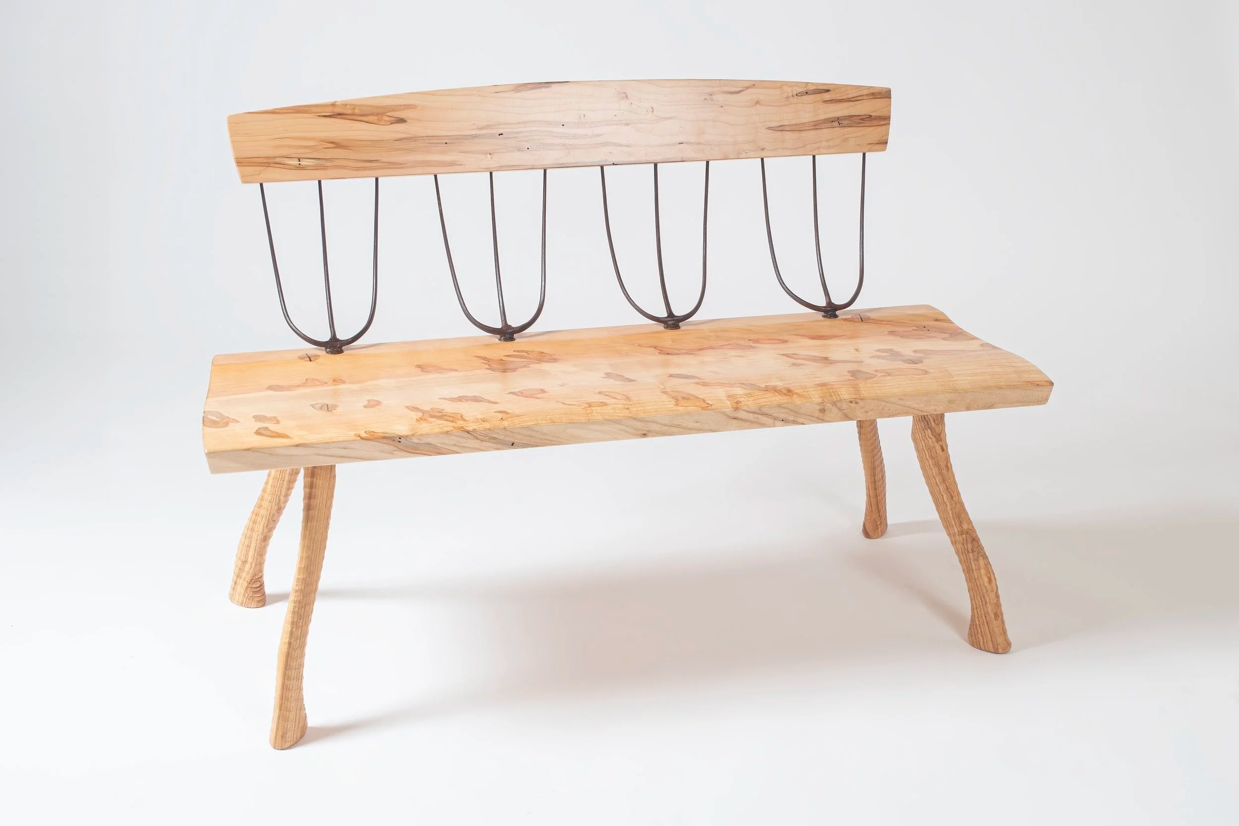 Handcrafted bench made from ambrosia maple with natural figuring and warm tonal variation. Four forged pitchfork heads serve as the back spindles, connecting the live-edge seat to a curved maple backrest. Legs are lathe-turned from ax handles with a natural outward splay. Photographed against a white background.