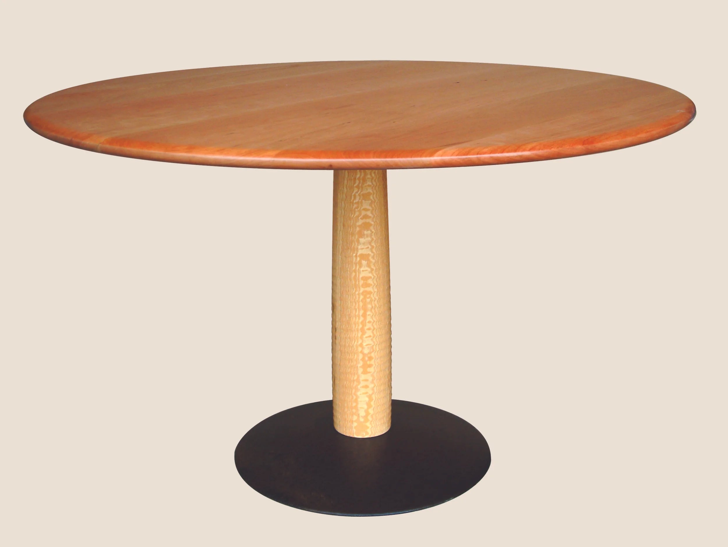 Round handcrafted pedestal dining table with a solid cherry top in a warm reddish amber tone and eased edge profile. The base is a single vintage lathe-turned ash post set into a flat recycled steel plow disc. Photographed against a neutral background.